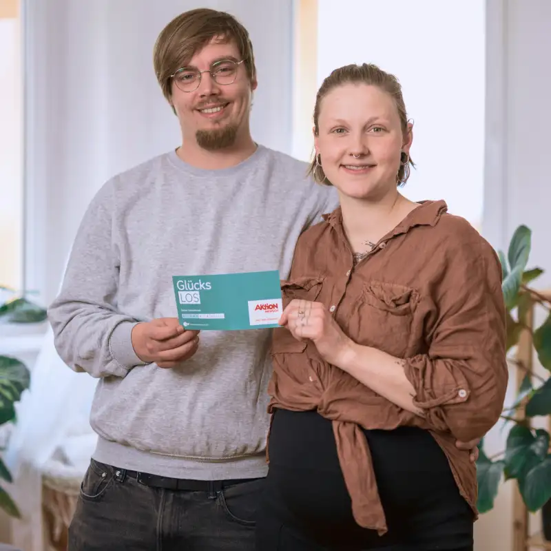 Aktion Mensch-Gewinner Sandro umarmt seine Freundin Fine. Sie stehen in ihrem Wohnzimmer und halten gemeinsam ein Aktion Mensch-Los fest.