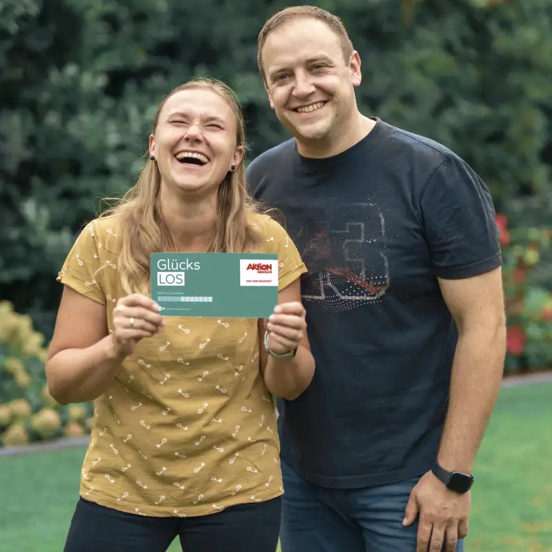 Aktion Mensch-Gewinnerin Bozena mit ihrem Mann im Garten. Sie lachen beide. Bozena hält ein Aktion Mensch-Los in der Hand.