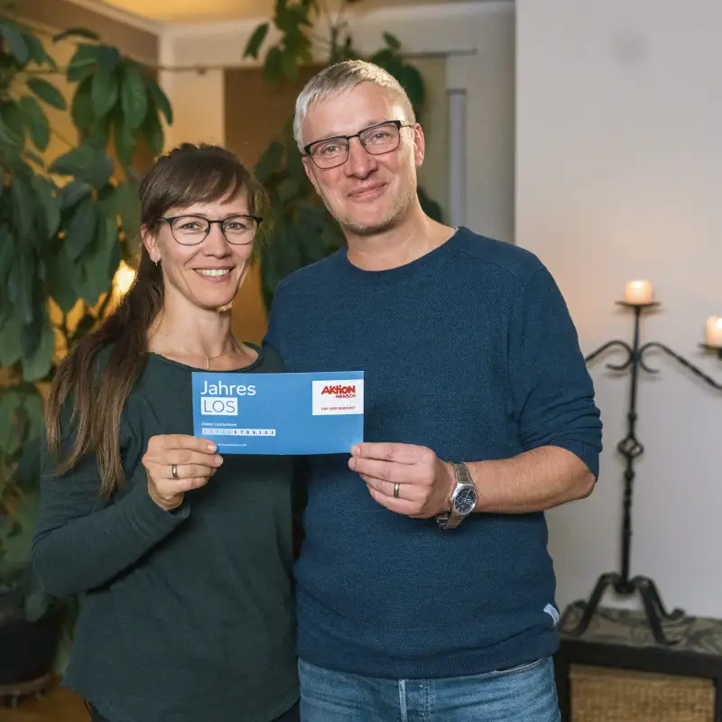 Aktion Mensch-Gewinner Jörg mit seiner Frau Adriane in ihrem Wohnzimmer. Gemeinsam halten sie ein Aktion Mensch-Los.