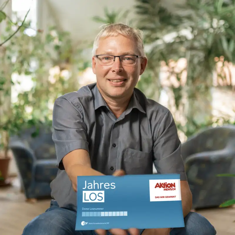Aktion Mensch-Gewinner Tobias sitzt in seinem Wohnzimmer. Er hält ein Aktion Mensch-Los in die Kamera.