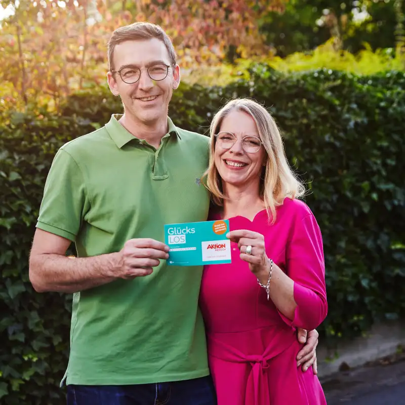 Aktion Mensch-Gewinner Stefan steht mit seiner Frau im Garten. Gemeinsam halten sie ein Aktion Mensch-Los fest.