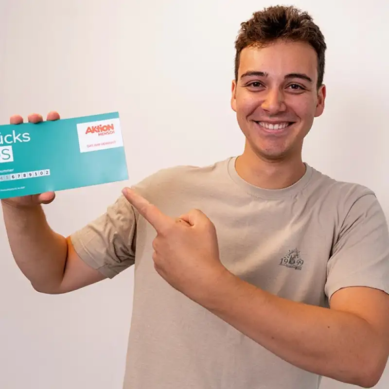 Aktion Mensch-Gewinner Benjamin steht vor einer hellen Wand. In der einen Hand hält er ein Aktion Mensch-Los mit der anderen Hand zeigt er auf dieses Los.