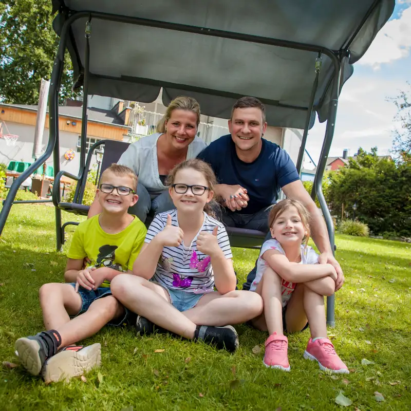 Aktion Mensch-Gewinner Marius mit Frau und Kindern im Garten. Die Eltern sitzen in einer Hollywood-Schaukel, die Kinder davor.