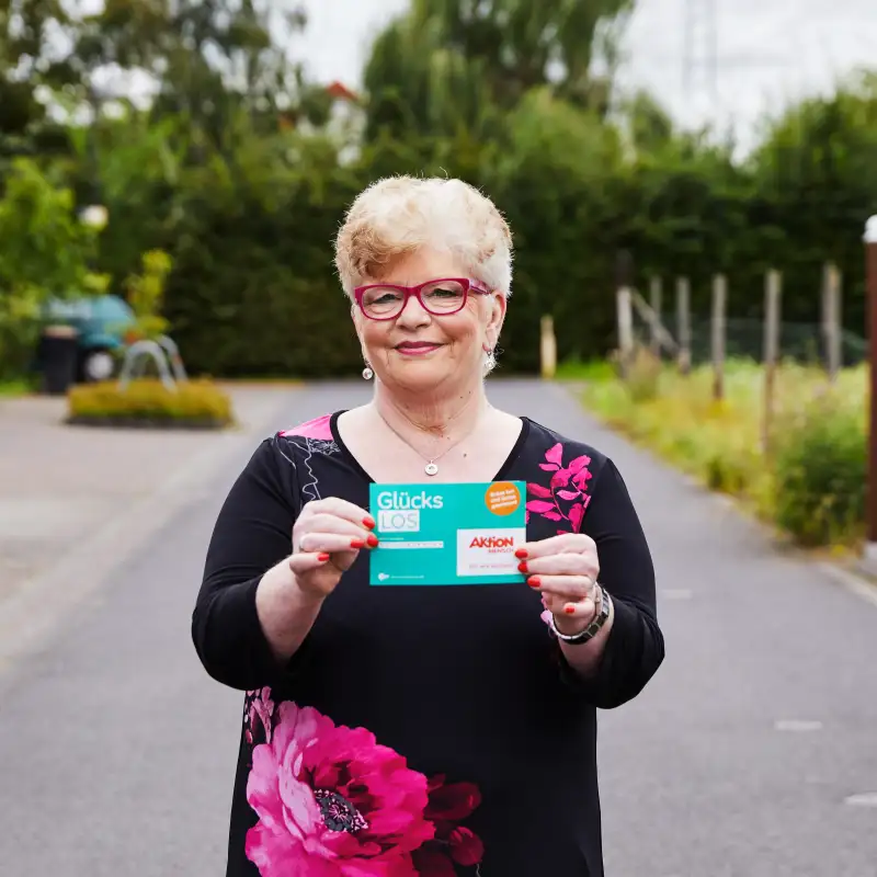 Aktion Mensch-Gewinner Gaby steht auf einer Straße. Sie hält ein Aktion Mensch-Los in der Hand