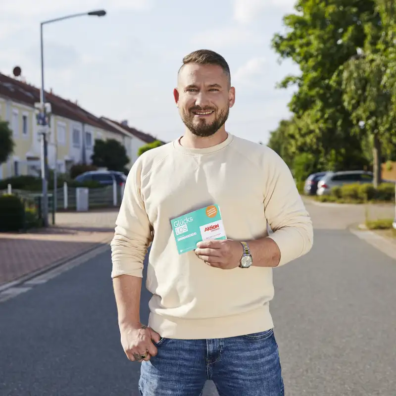 Aktion Mensch-Gewinner Anton steht auf einer Straße und hält ein Aktion Mensch-Los in der Hand.