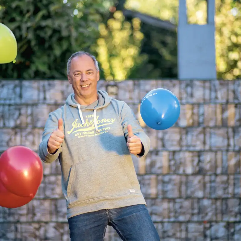 Aktion Mensch-Gewinner Jürgen steht in seinem Garten. Er reckt beide Daumen hoch und es fliegen Luftballons.