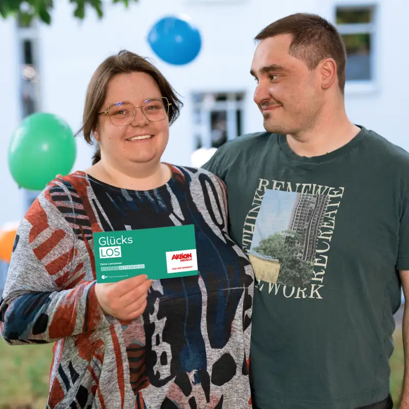 Aktion Mensch-Gewinner Felix und seine Frau Luisa vor einem Haus. Luisa hält ein Aktion Mensch-Los in der Hand. Im Hintergrund schweben Luftballons.