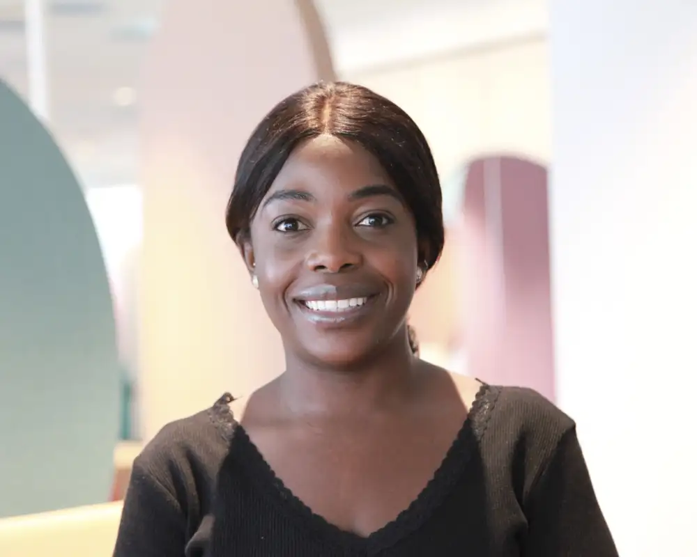Benedicte Mayabu, eine junge Frau mit langen dunklen Haaren steht in einem Aktion Mensch-Büro und lächelt in die Kamera.