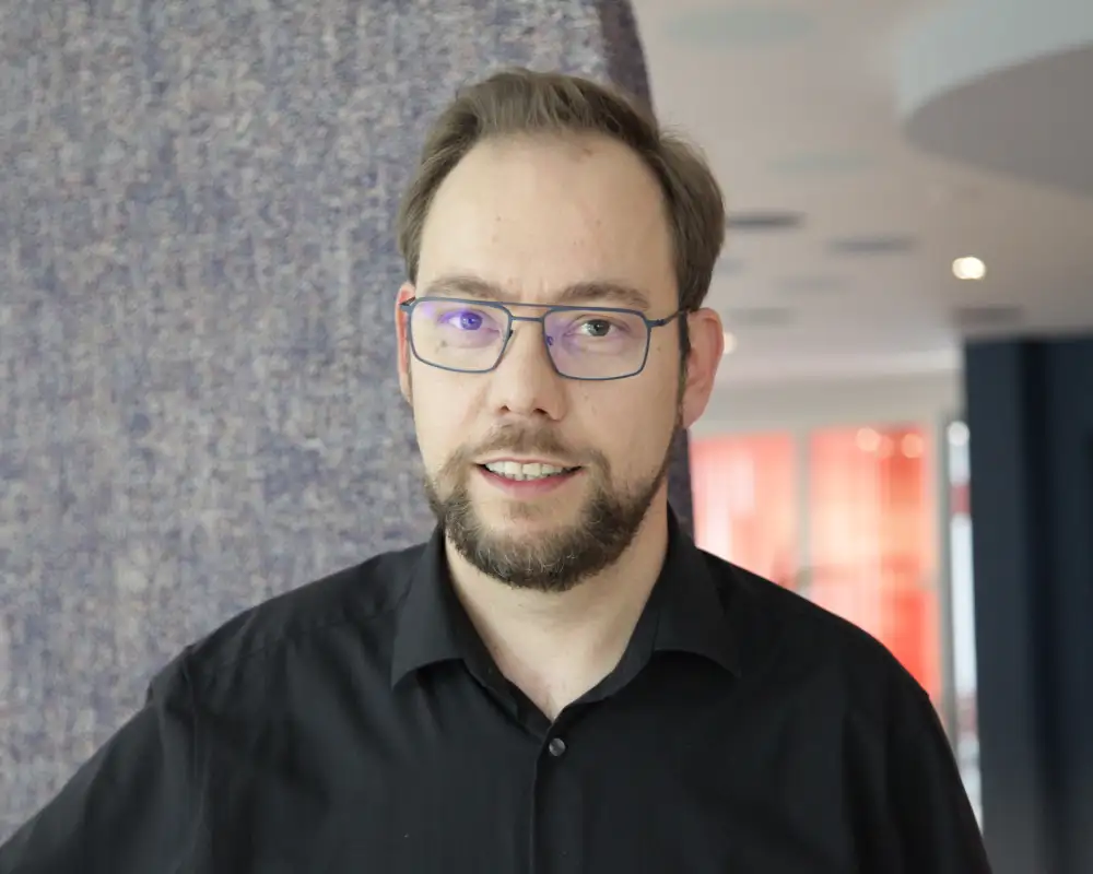 Alexander Westheide - ein Mann mit kurzen braunen Haaren, Bart, Schnäuzer und Brille steht in einem Aktion Mensch-Büro.