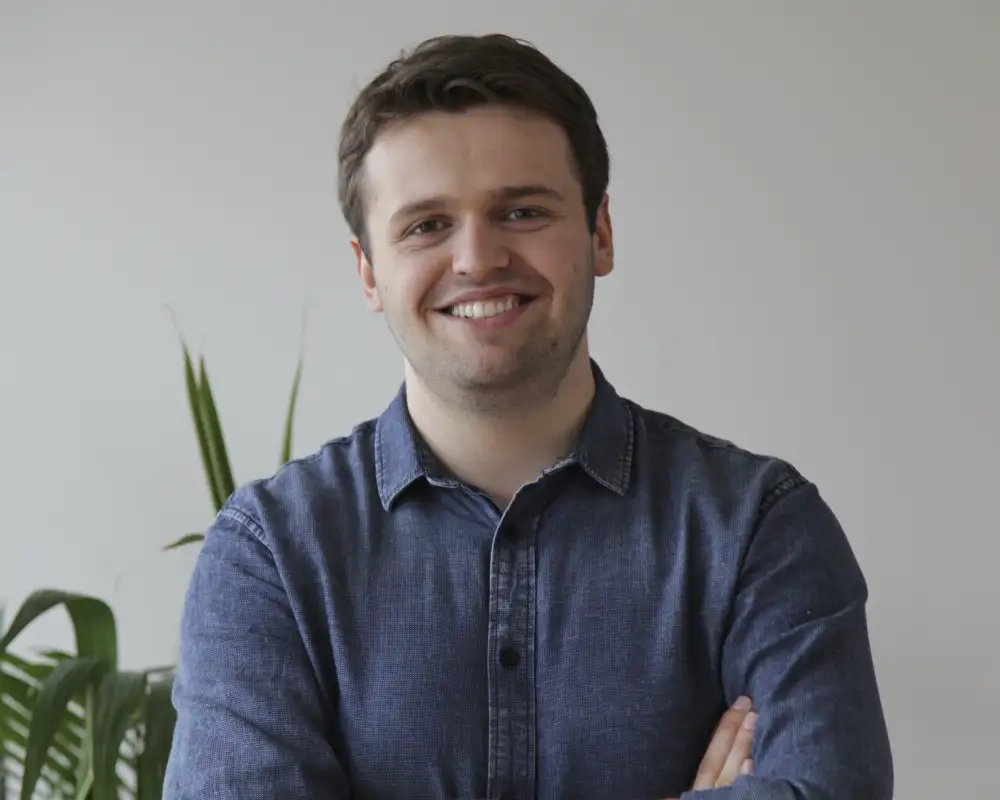 Eric Merten, ein junger Mann mit kurzen braunen Haaren steht mit verschränkten Armen vor einer weißen Wand und lächelt in die Kamera.