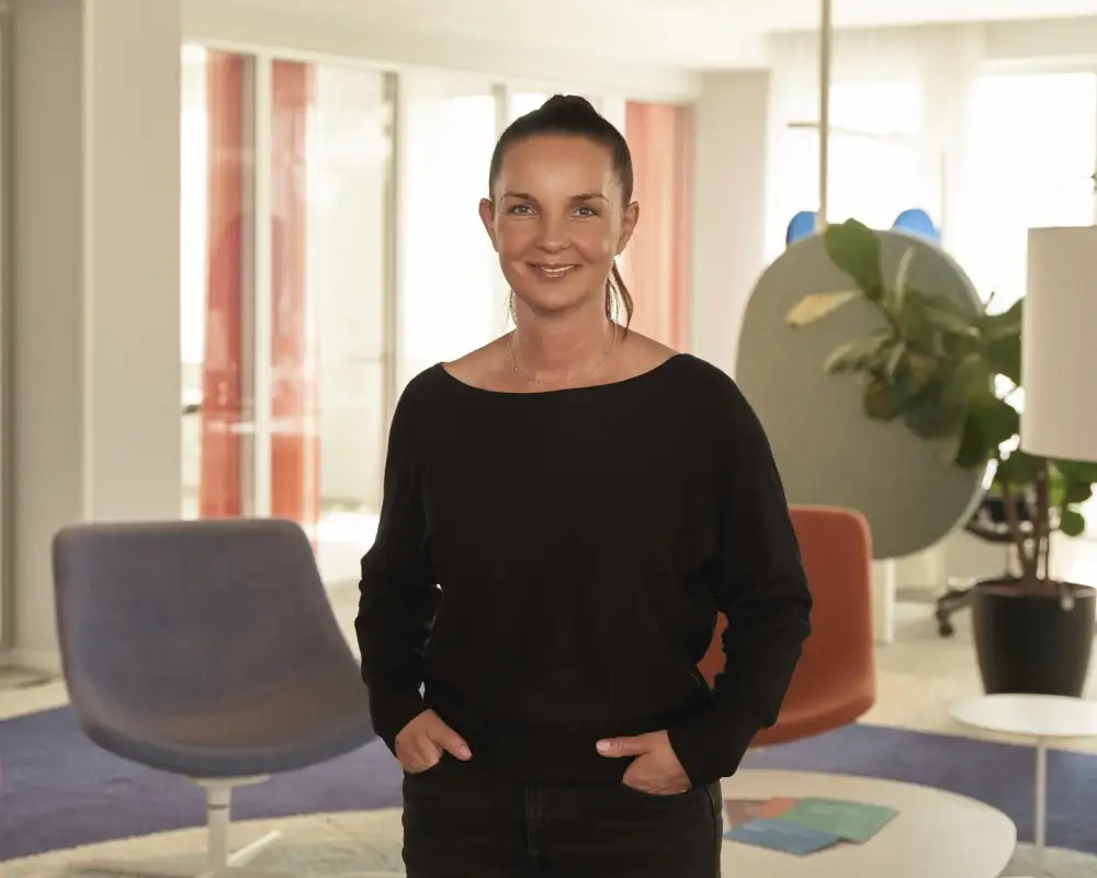 Jana Stollenwerk - eine Frau mit langen dunklen Haaren, die zu einem Pferdeschwanz gebunden sind. Sie steht mit den Händen in den Hosentaschen in einem Aktion Mensch-Büro.