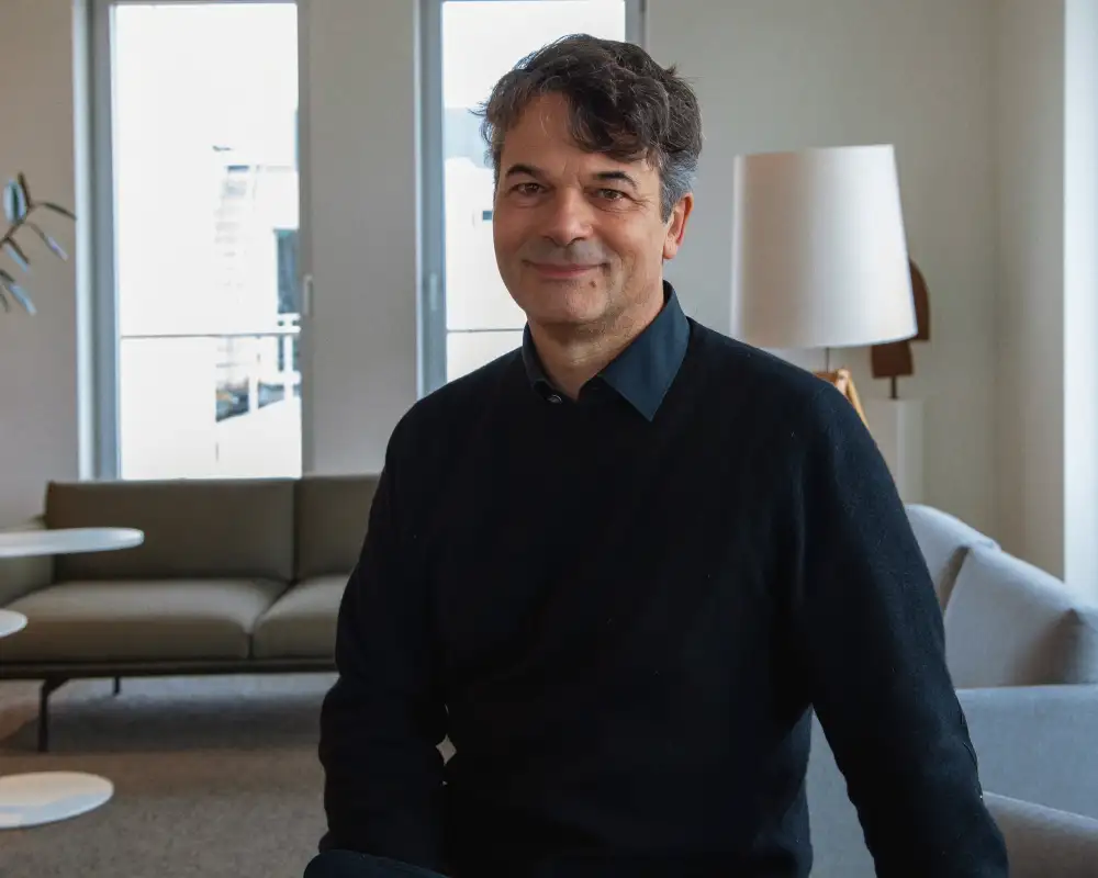 Robert trägt einen schwarzen Pullover und ein schwarzes Hemd. Er steht vor einem Sofa im Aufenthaltsraum und lächelt in die Kamera.