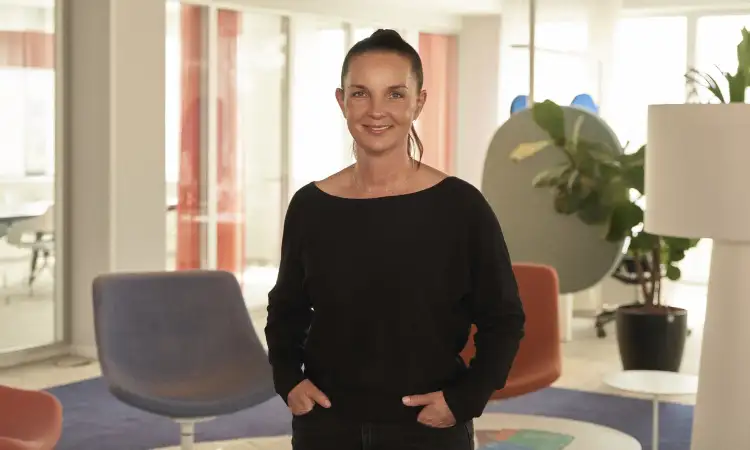 Jana Stollenwerk - eine Frau mit langen dunklen Haaren, die zu einem Pferdeschwanz gebunden sind. Sie steht mit den Händen in den Hosentaschen in einem Aktion Mensch-Büro.