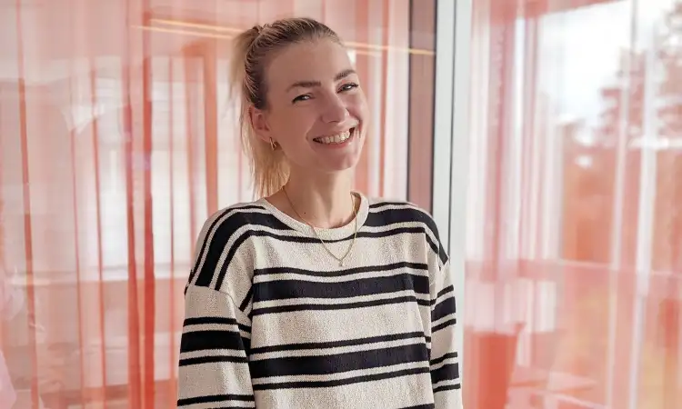 Halbtotale von Marie Molitor. Einer jungen Frau in einem schwarz-weiß gestreiften Pulli, die vor einer Glaswand mit Vorhängen steht. Sie lacht in die Kamera. Ihre langen blonden Haare sind zu einem Pferdeschwanz gebunden.