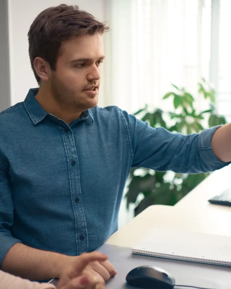 Ein junger Mann und eine Frau sitzen nebeneinander vor einem Bildschirm. Der junge Mann zeigt mit der linken Hand auf den Monitor.
