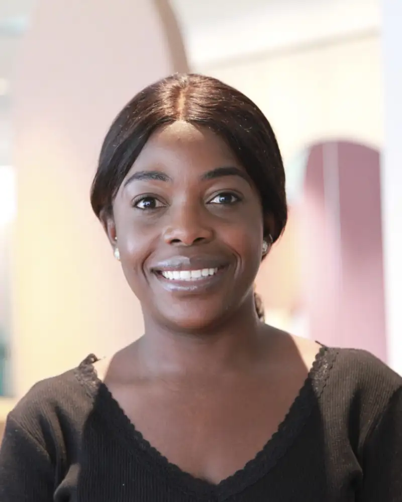 Benedicte Mayabu, eine junge Frau mit langen dunklen Haaren steht in einem Aktion Mensch-Büro und lächelt in die Kamera.