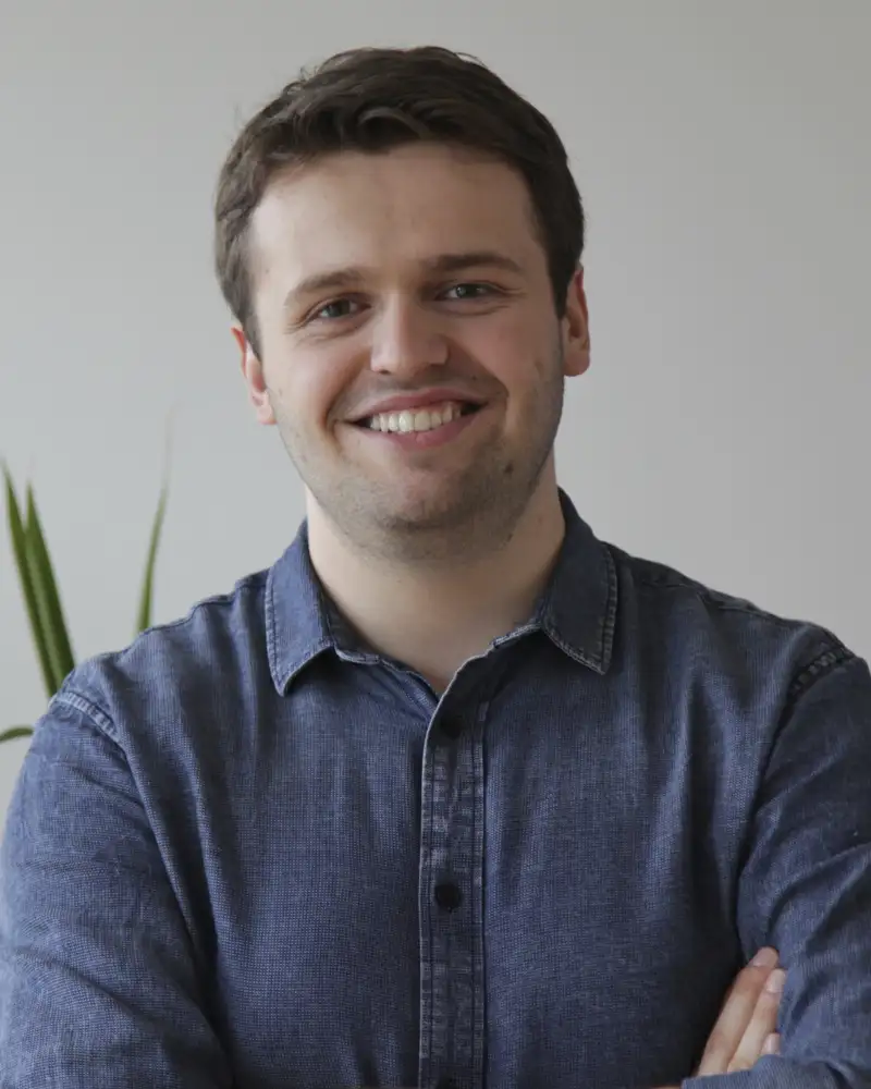 Eric Merten, ein junger Mann mit kurzen braunen Haaren steht mit verschränkten Armen vor einer weißen Wand und lächelt in die Kamera.