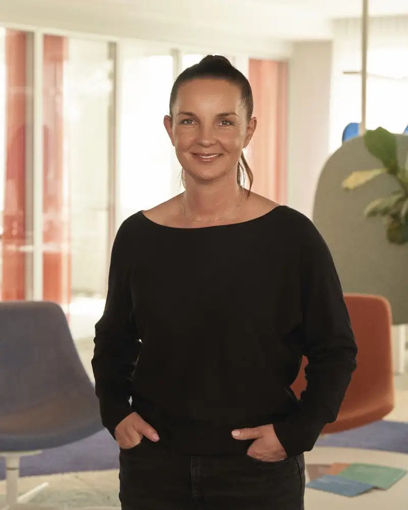 Jana Stollenwerk - eine Frau mit langen dunklen Haaren, die zu einem Pferdeschwanz gebunden sind. Sie steht mit den Händen in den Hosentaschen in einem Aktion Mensch-Büro.