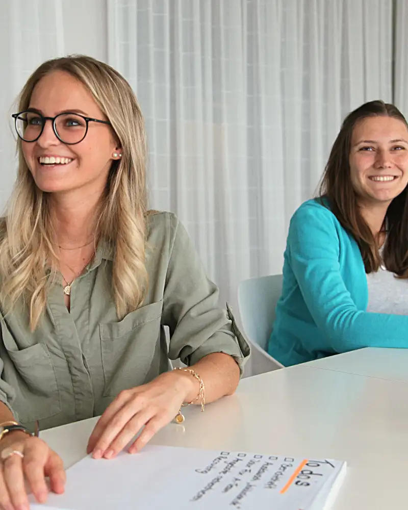 Zwei junge Frauen sitzen nebeneinander an einem großen Tisch in einem Konferenzraum. Die Vordere der beiden hat einen Block vor sich auf dem Tisch liegen. Dort ist die Überschrift "To Dos" zu lesen. Beide Frauen lachen freundlich.