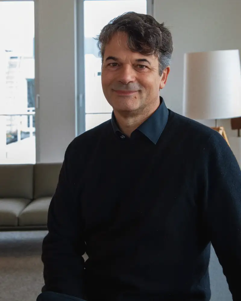 Robert trägt einen schwarzen Pullover und ein schwarzes Hemd. Er steht vor einem Sofa im Aufenthaltsraum und lächelt in die Kamera.