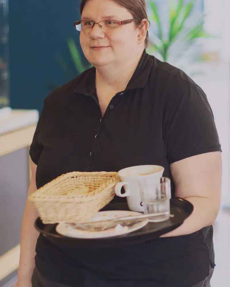 Eine Frau in einem Café. Sie trägt ein Tablett mit Frühstücksgeschirr.
