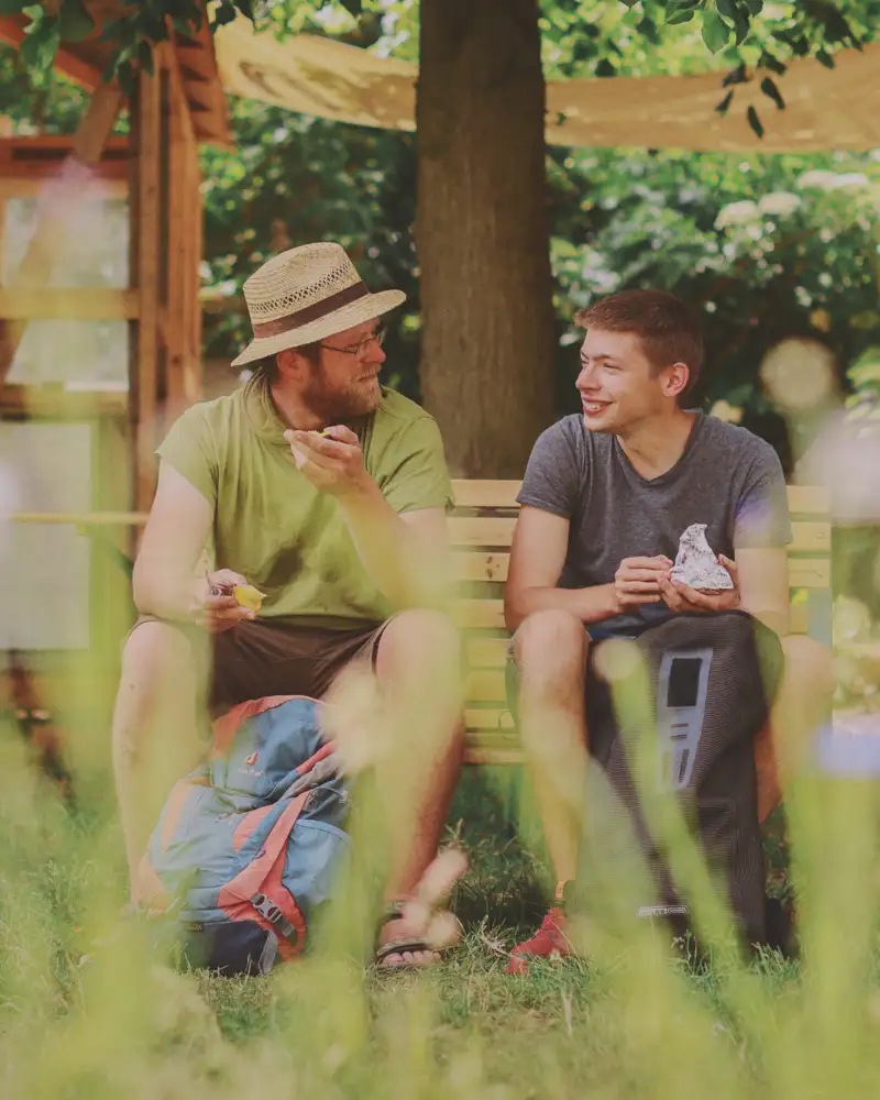 Zwei Männer, einer mit Strohhut, sitzen in einem Garten auf einer Bank und unterhalten sich.