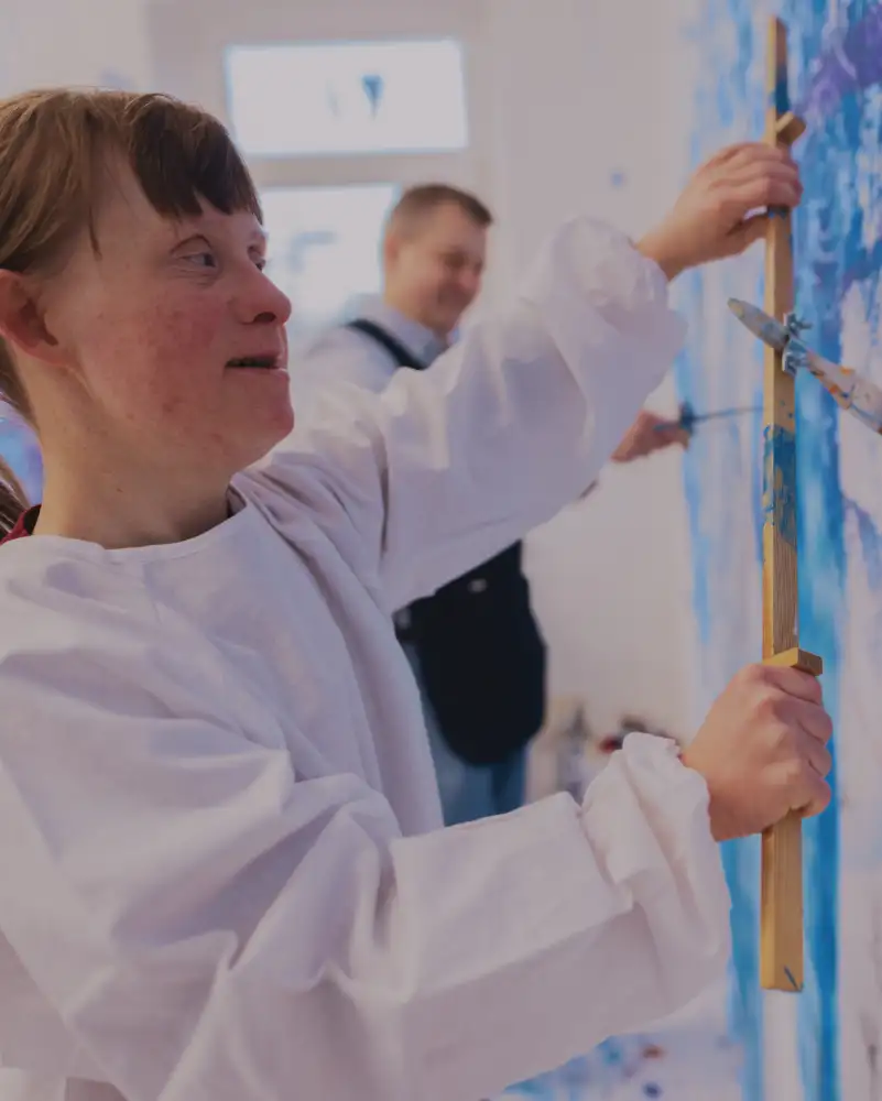 Eine Frau mit Downsyndrom im weißen Kittel steht vor einer großen weißen, blau bemalten Leinwand.