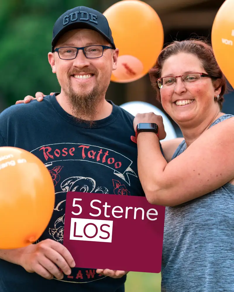 Ein Mann und eine Frau stehen im Garten. Die Frau umarmt den Mann. Der Mann hält ein 5 Sterne Los in der Hand. Beide lachen und freuen sich.
