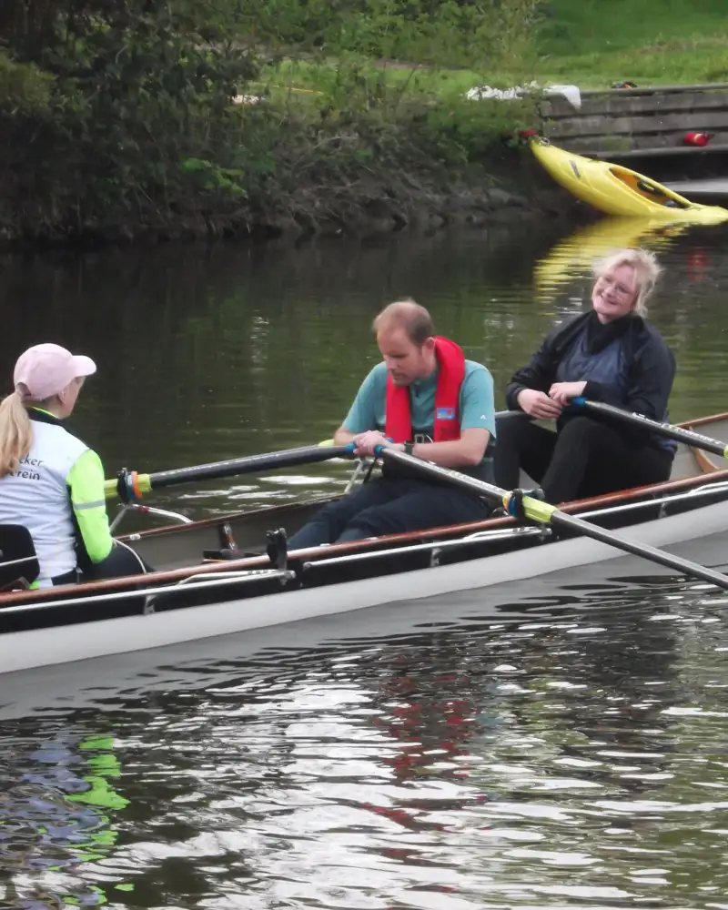Drei Personen sitzen auf einem Gewässer in einem Ruderboot unterwegs.