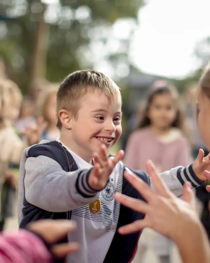 Eine Gruppe Kinder aus der Integrativen Kita Blumenwiese in Grevenbroich schaut auf einen Jungen mit Down-Syndrom der fröhlich auf eine Frau zuläuft.