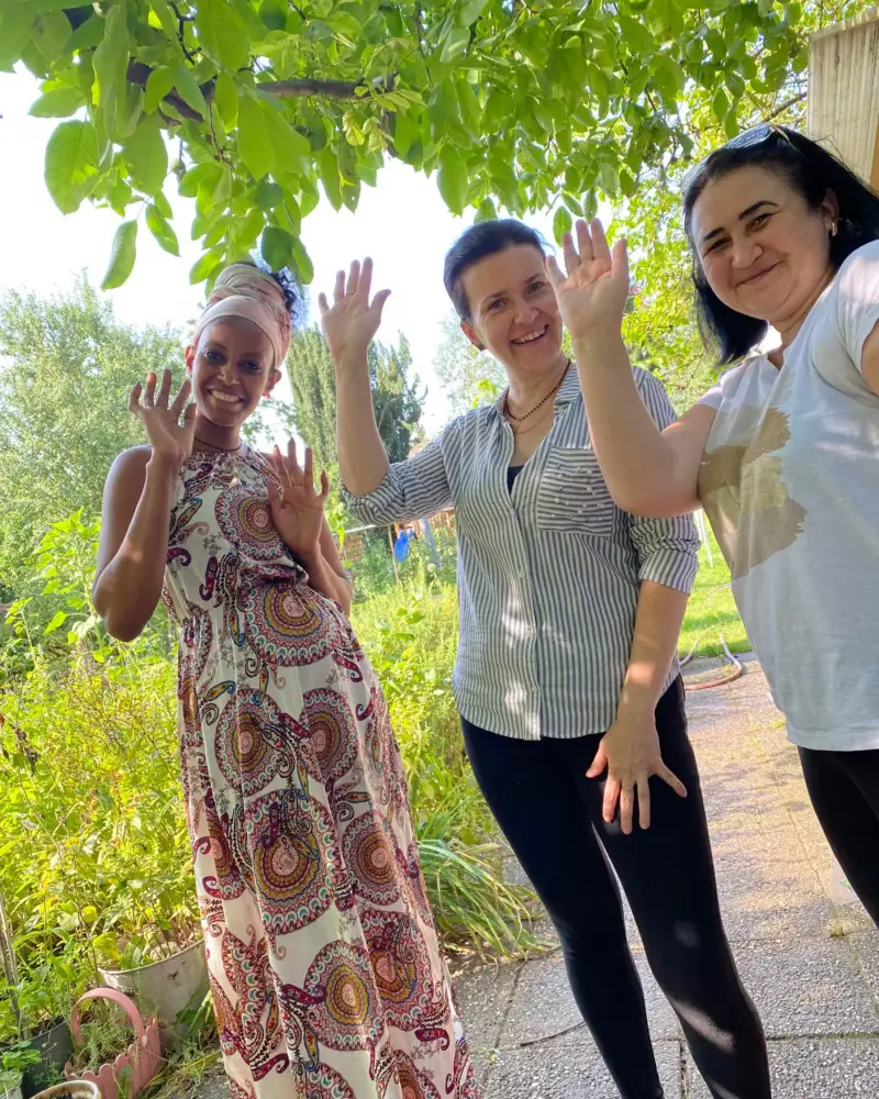 Drei Frauen stehen in einem Garten und winken in die Kamera.