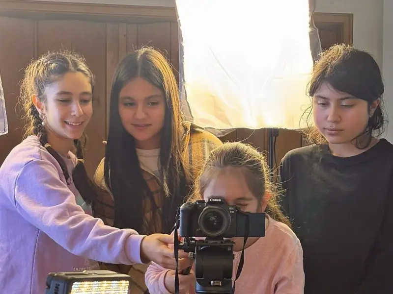 Eine Gruppe von vier Mädchen. Eines von ihnen schaut durch die Linse einer Kamera, die anderen sehen dabei zu.