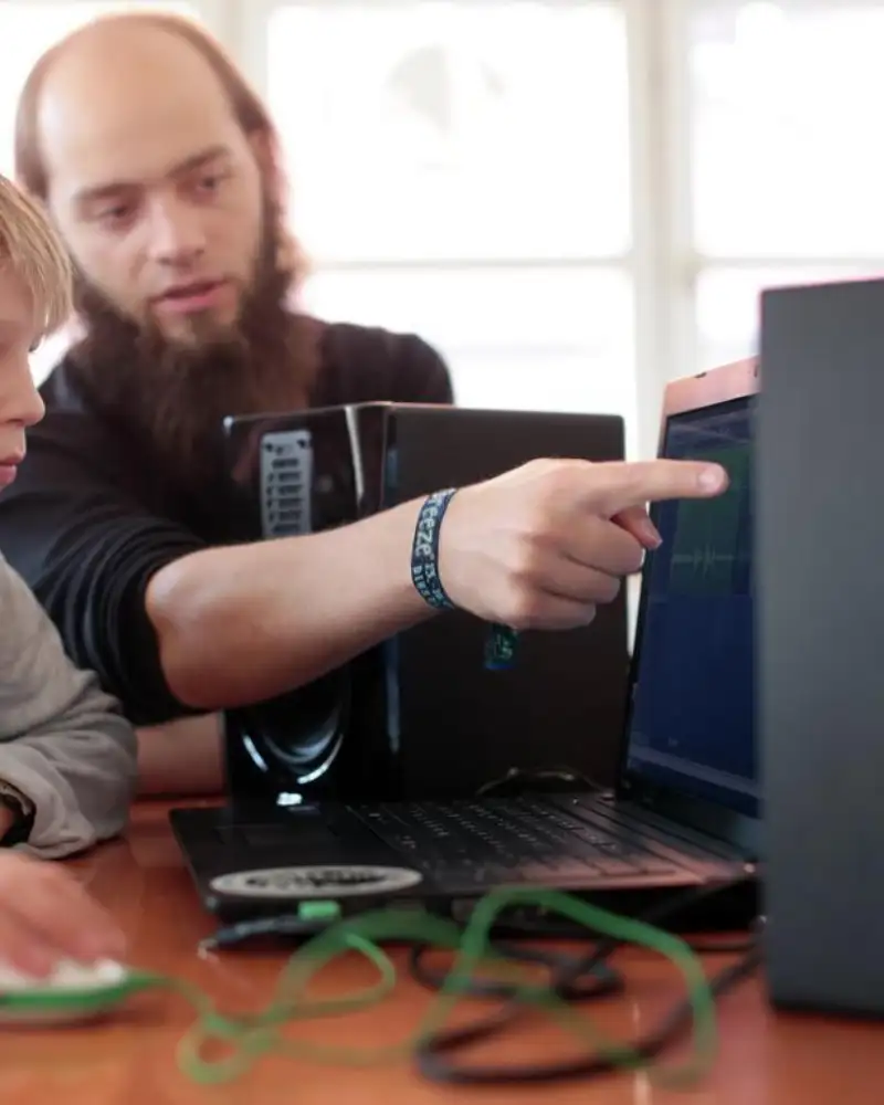 Ein Junge und ein junger Erwachsener sitzen vor einem Laptop. Der junge Erwachsene zeigt mit dem Finger auf den Bildschirm.
