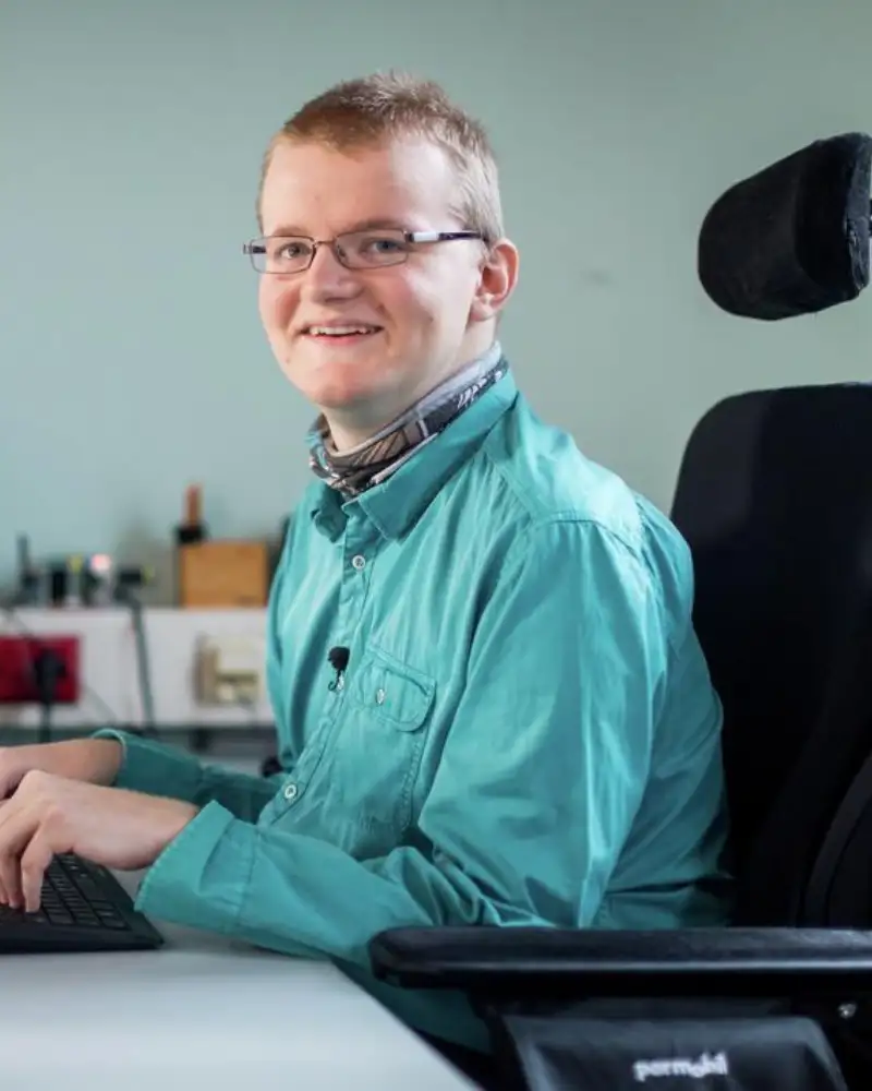 Ein junger Mann sitzt im Rollstuhl am Schreibtisch. Seine Hände liegen auf der Tastatur, er lächelt in die Kamera.