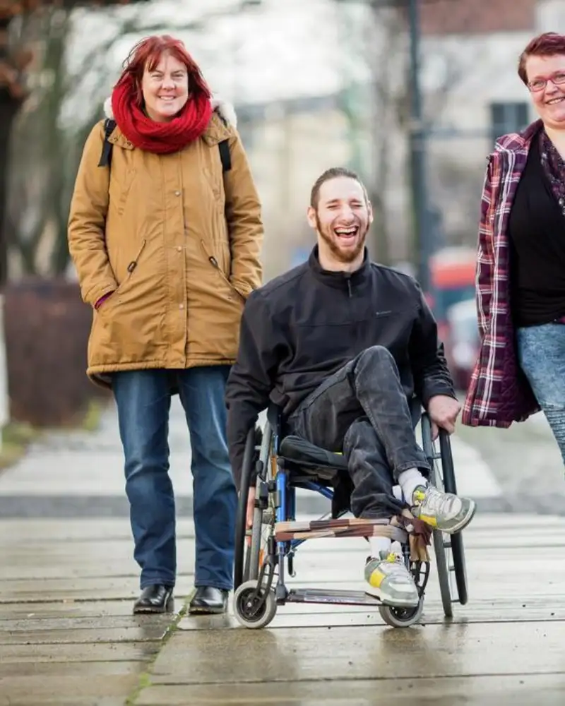 Drei junge Menschen stehen neben einander auf dem Bürgersteig. Einer von ihnen sitzt im Rollstuhl.