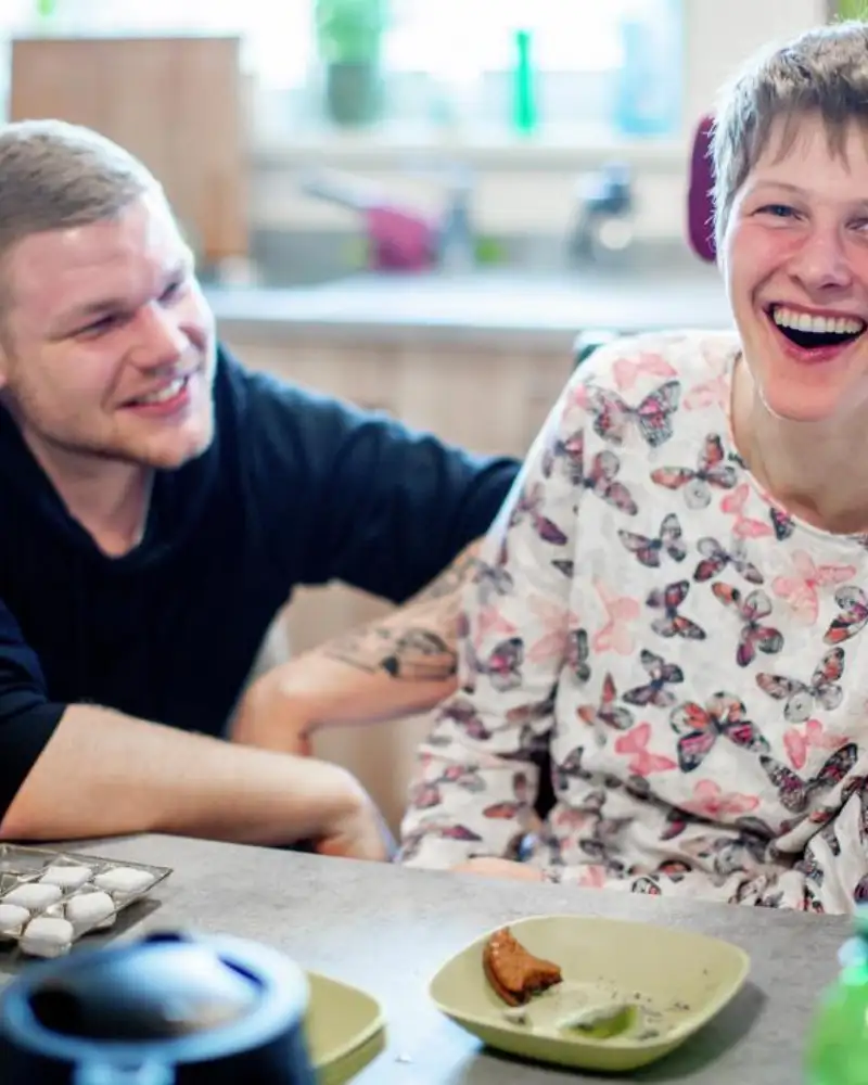 Zwei junge Menschen an einem Tisch. Die junge Frau sitzt im Rollstuhl. Der junge Mann hockt neben ihr.
