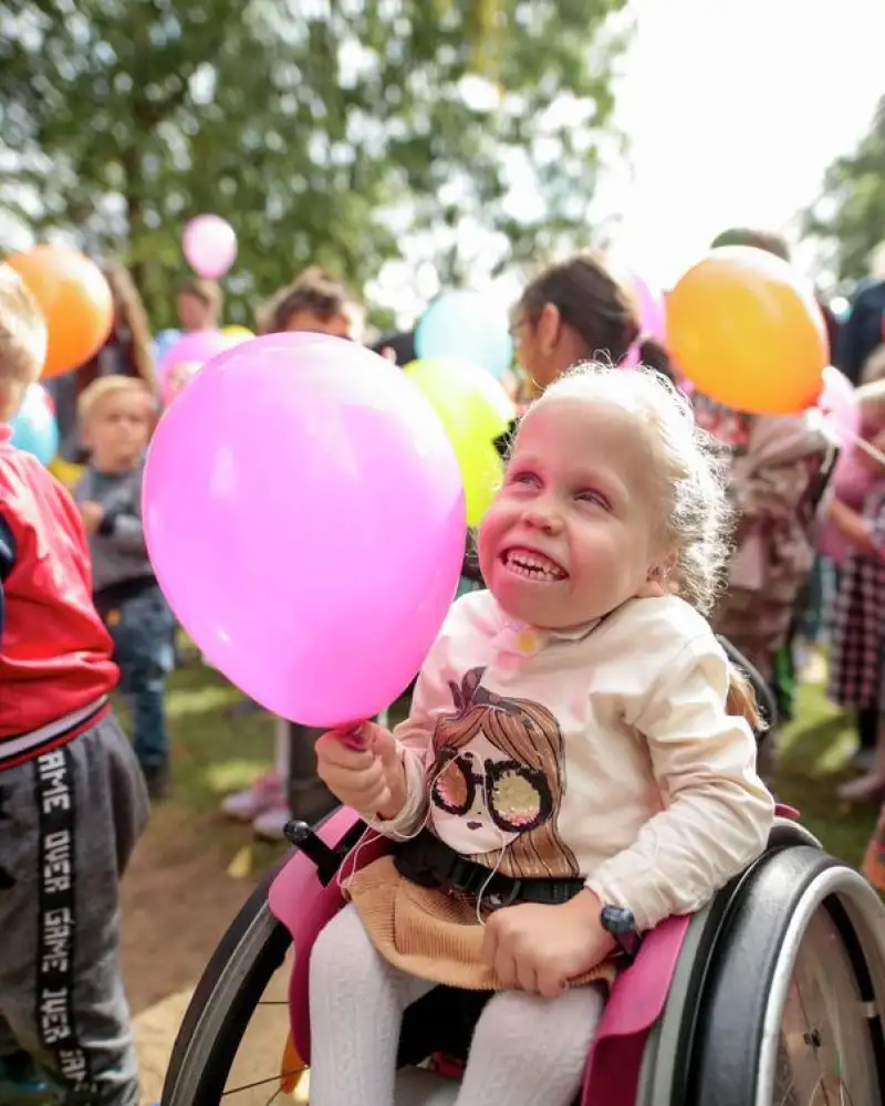 Mehrere Kleinkinder draußen mit Luftballons. Das Mädchen im Vordergrund sitzt im Rollstuhl.