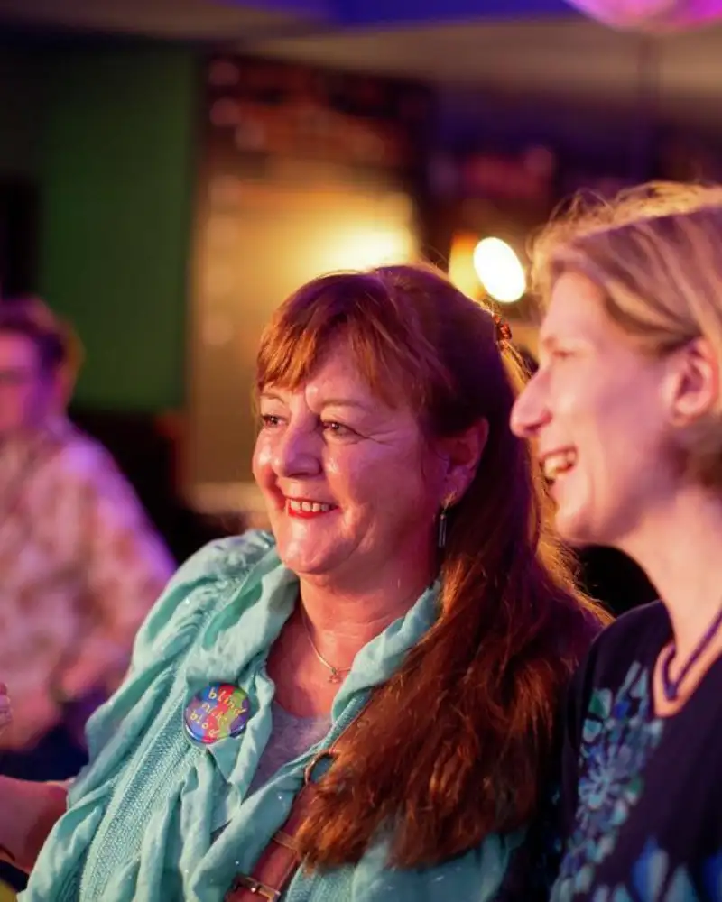Zwei Frauen sitzen nebeneinander in einem dunklen Saal und schauen nach vorne. Sie lachen, offensichtlich erheitert sie das, was sie dort sehen.