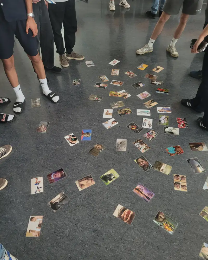 Jugendliche, von denen man nur die Beine sieht, stehen um viele Fotos herum, die vor ihnen auf dem Boden ausgebreitet sind.