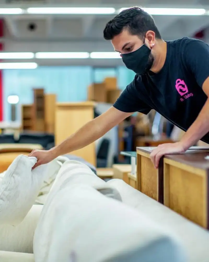 Ein junger Mann beugt sich in einem Kaufhaus von rechts über ein Sofa und hat ein Kissen in der Hand.