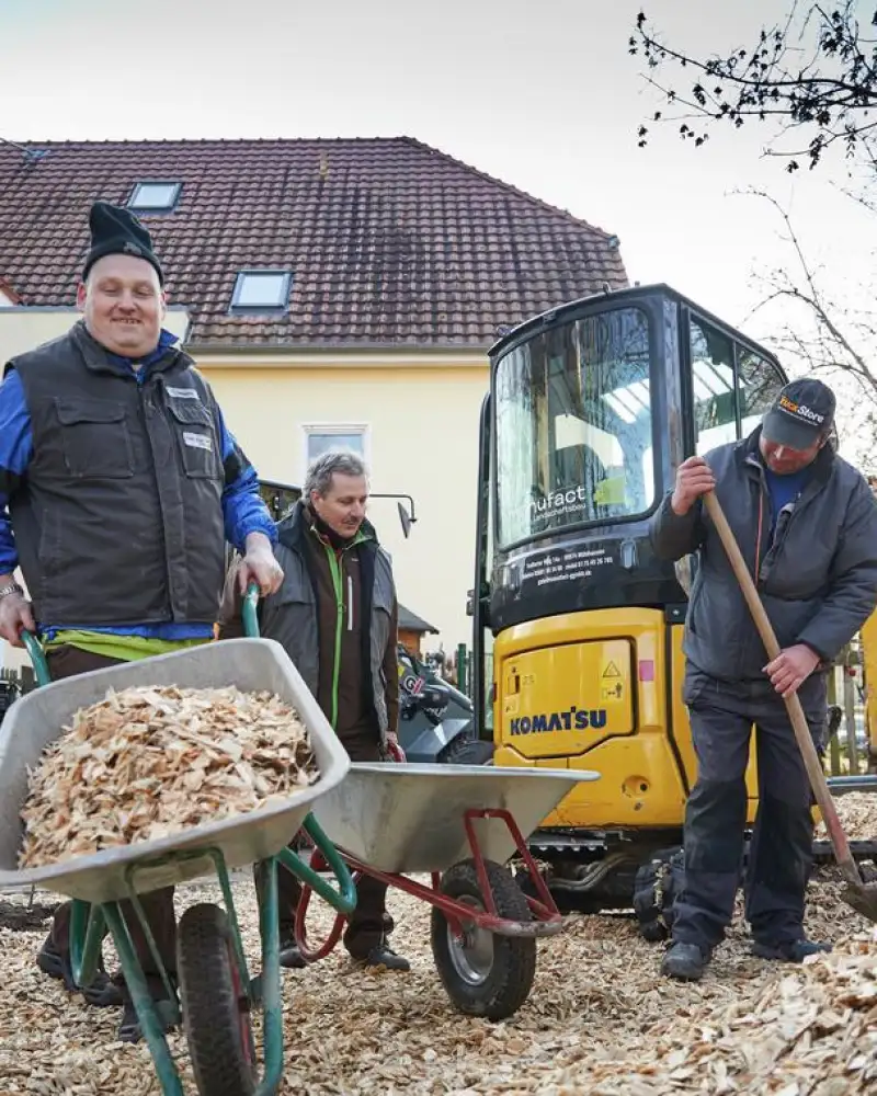 Zwei Männer mit Schubkarre und ein Mann mit Spaten. Im Hintergrund steht ein Bagger.