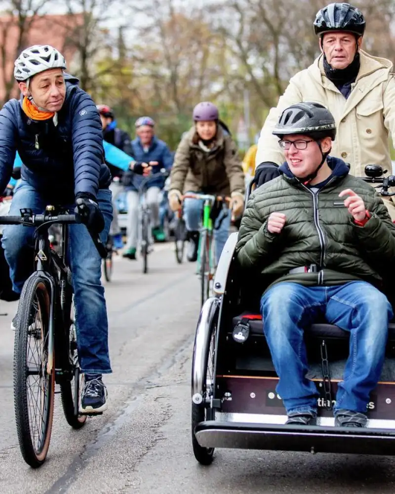 Vorneweg eine Fahrradrikscha mit einem Fahrer und einem Fahrgast. Daneben und dahinter ist eine Gruppe von Radlern unterwegs.