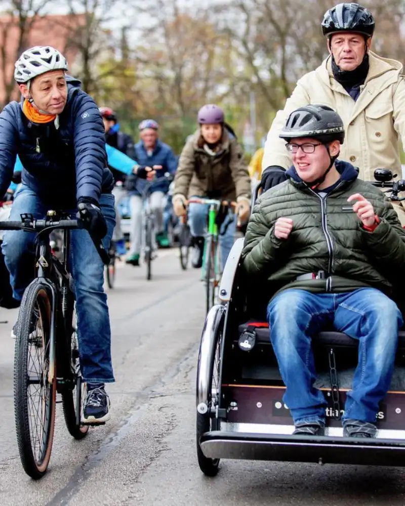 Vorneweg eine Fahrradrikscha mit einem Fahrer und einem Fahrgast. Daneben und dahinter ist eine Gruppe von Radlern unterwegs.