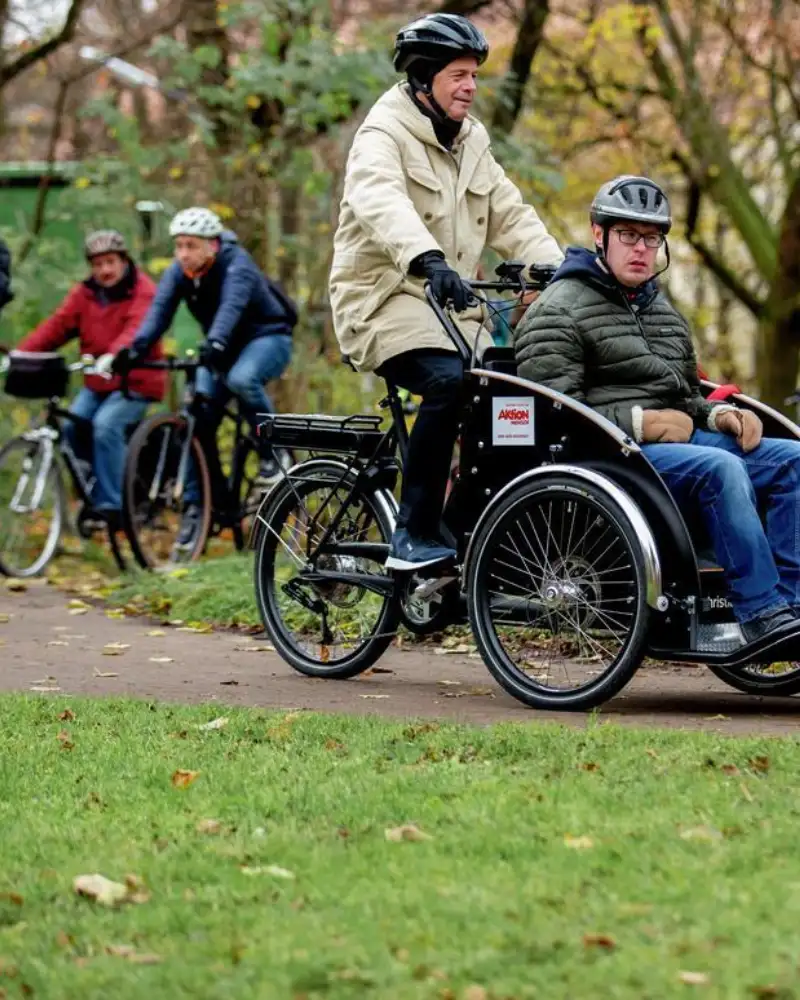 In einem Park fährt vorneweg eine Fahrradrikscha mit einem Fahrer und einem Fahrgast. Dahinter nähert sich eine Gruppe von Radlern.