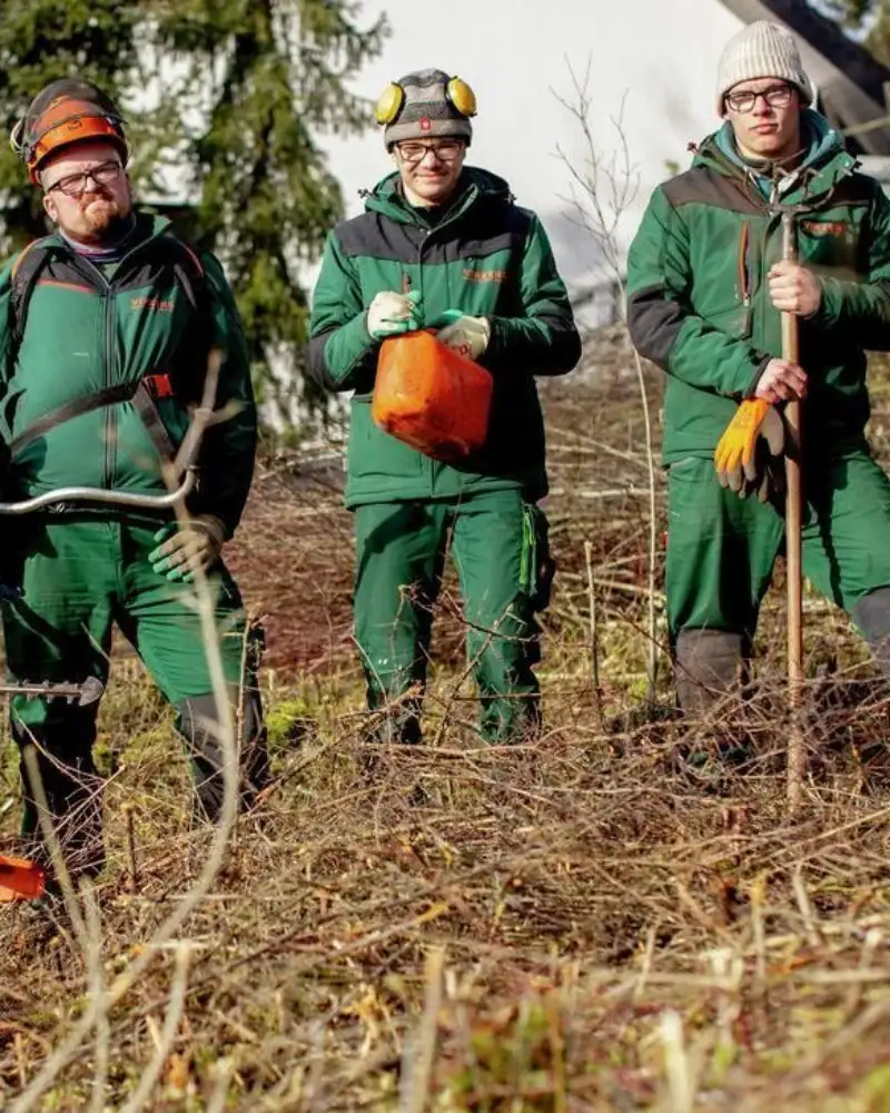 Drei Männer in grüner Arbeitskleidung mit Gartengeräten stehen an einem Hang voller Gestrüpp. Zwei von ihnen tragen Helme und Kopfhörer, die anderen beiden Mützen.