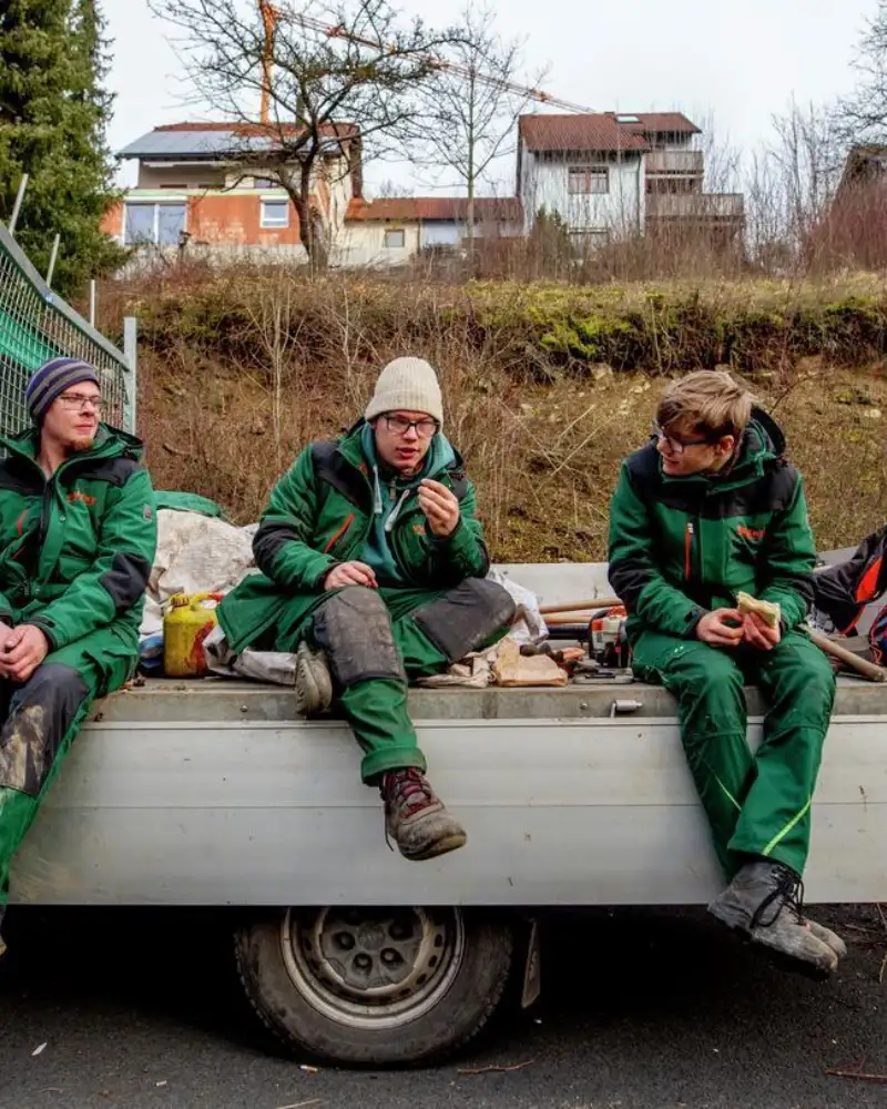Drei junge Männer in grüner Arbeitskleidung machen Pause. Dabei sitzen auf der Ladefläche eines Transporters.