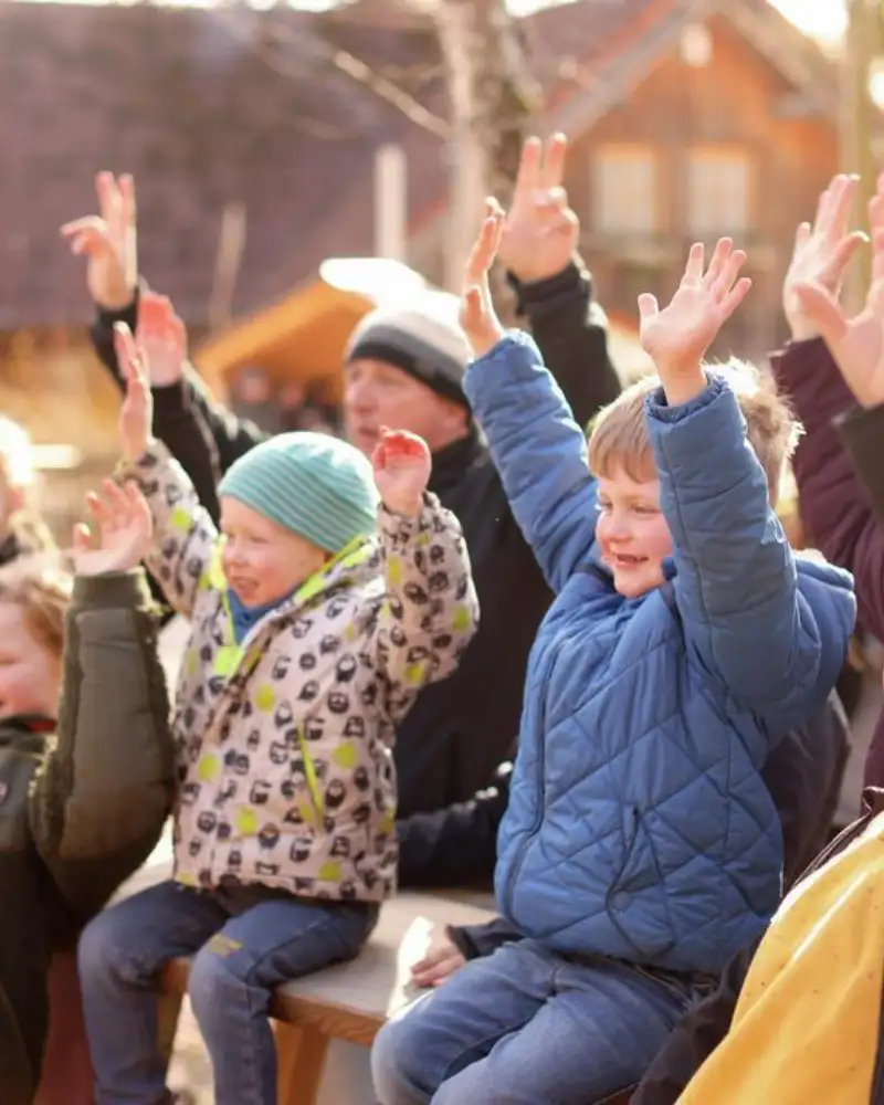 Mehrere Kinder und Erwachsene sitzen in Jacken draußen. Sie reißen ihre Arme hoch und jubeln.