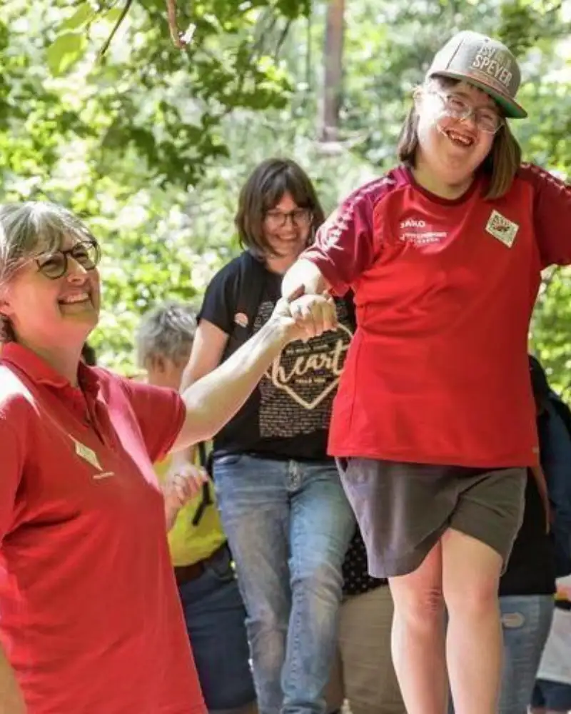 Ein Mädchen mit Down-Syndrom balanciert über einen Baumstamm. Dabei hält eine Frau ihre Hand. Dahinter folgt ein weiteres Mädchen. Weitere Personen stehen dabei.