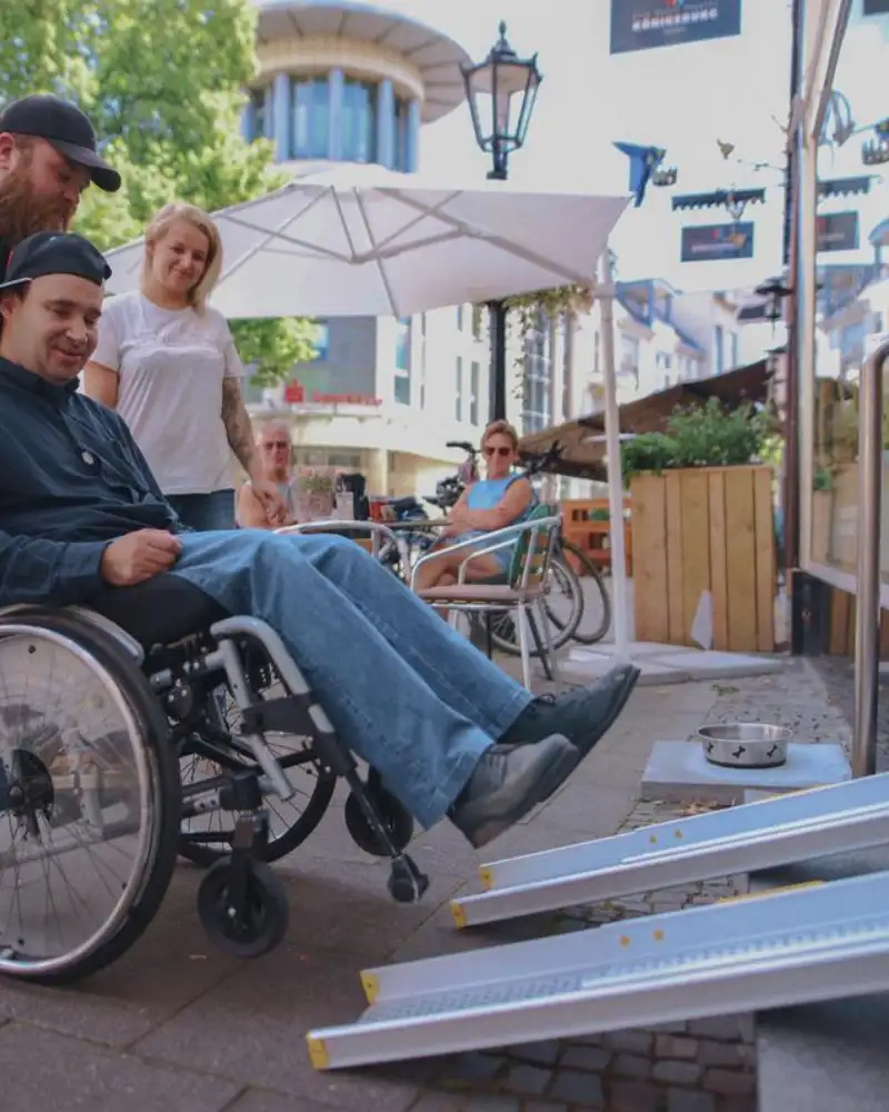 Ein Mann hilft einem anderen jungen Mann im Rollstuhl eine Rampe hinauf. Eine junge Frau schaut dabei zu.