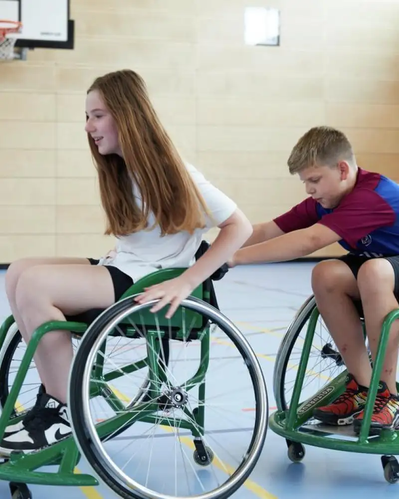 Zwei Kinder in einer Turnhalle im Rollstuhl. Das Mädchen ist vorne, der Junge hinter ihr. Er hält sich an ihrem Rollstuhl fest.
